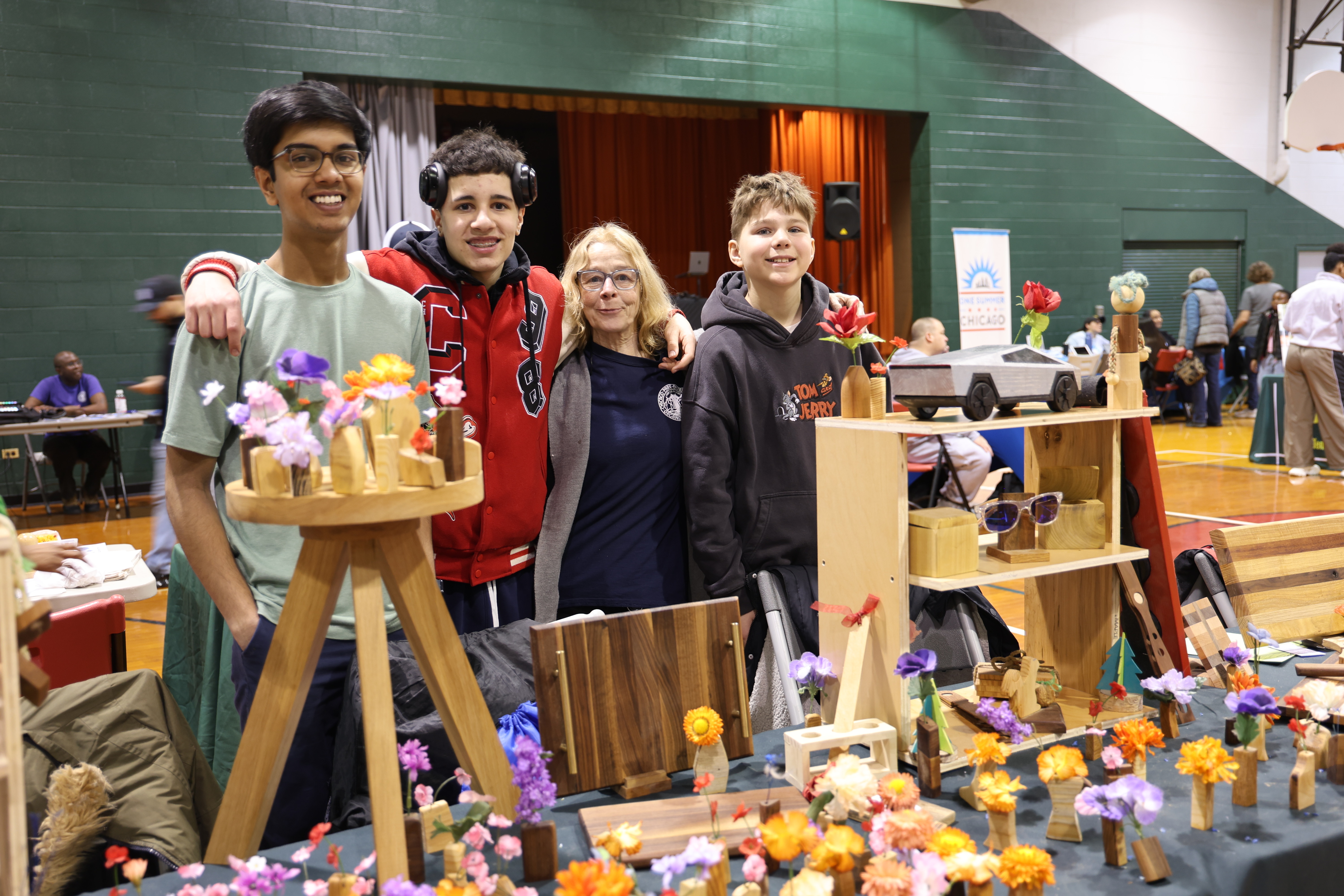 Four smiling people pose with a display of small wooden crafts and colorful artificial flowers.